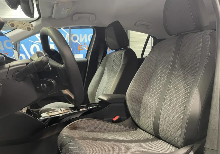 Vue intérieure côté conducteur de la Peugeot 208 1.2i Hybrid 2025 avec sièges tissu gris et accoudoir central noir.