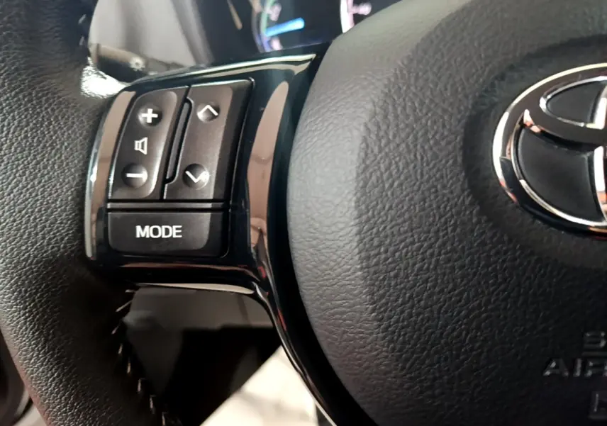 Gros plan sur les commandes gauche du volant en cuir noir de la Toyota Yaris hybride blanche, avec logo Toyota visible.