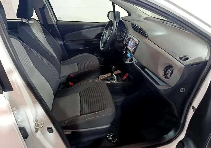 Vue intérieure côté conducteur de la Toyota Yaris blanche 2019, montrant sièges tissu bicolore et tableau de bord avec écran tactile.