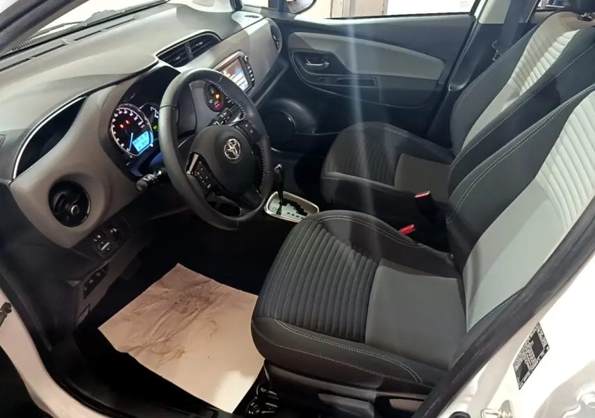 Vue intérieure côté conducteur de la Toyota Yaris blanche 2019, montrant sièges bi-ton et tableau de bord avec écran tactile.