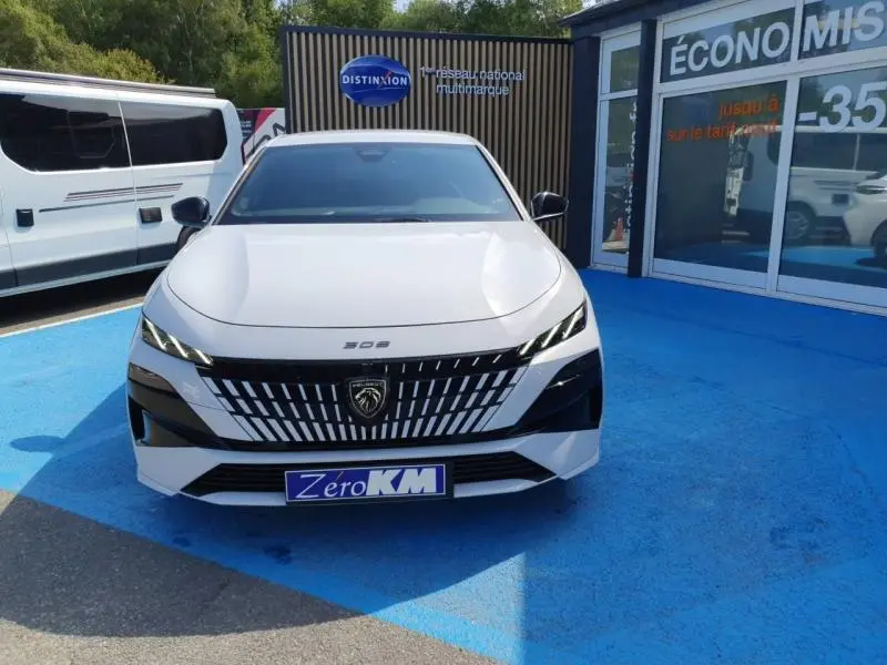 Vue avant d'une Peugeot 308 blanche Okénite avec calandre distinctive et phares LED fins, stationnée sur sol bleu.