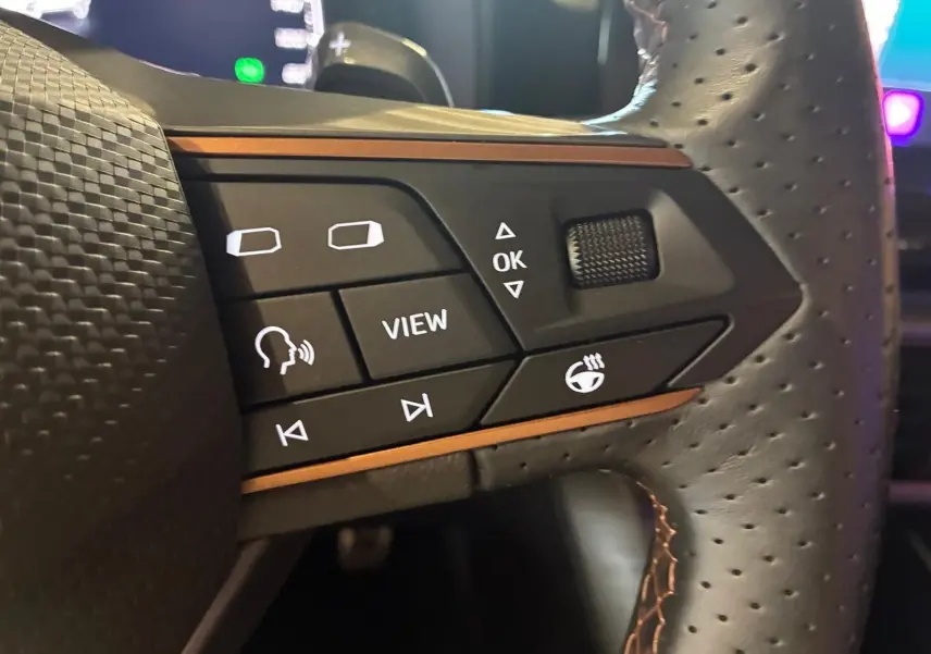 Gros plan sur les commandes du volant perforé du CUPRA Formentor 1.4 E-HYBRID 2024, avec surpiqûres orange et boutons multifonctions.