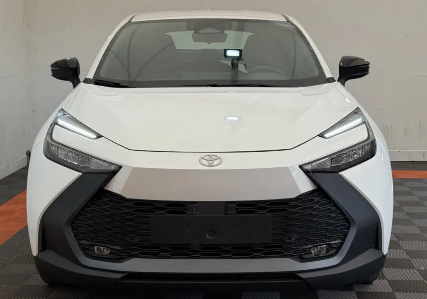 Vue frontale d'un Toyota C-HR 1.8 Hybride blanc avec calandre noire et phares LED fins, en intérieur sur sol damier.