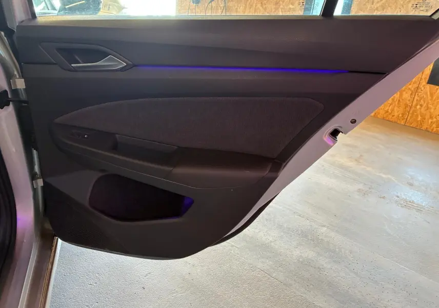 Vue rapprochée de la porte arrière gauche intérieure d’une Volkswagen Golf gris clair 2023 avec éclairage d’ambiance bleu.