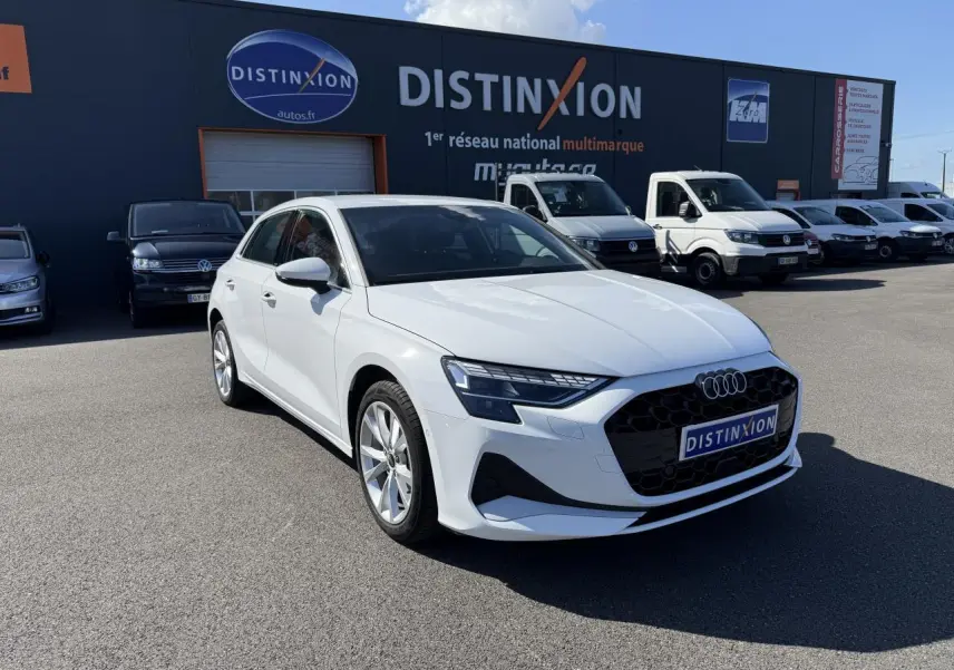 Audi A3 Sportback blanc Arcona vue 3/4 avant droit sur parking avec bâtiment Distinxion en arrière-plan.
