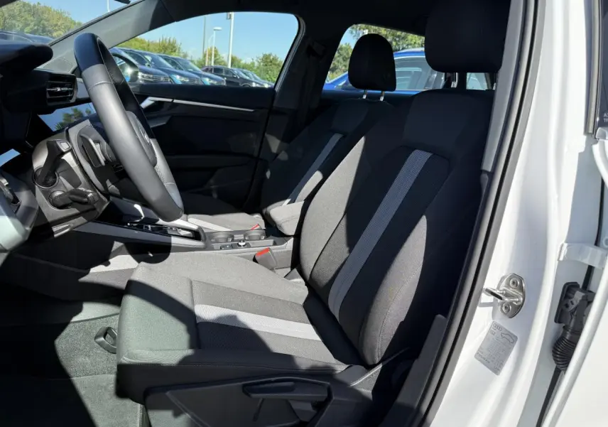 Intérieur côté conducteur de l'Audi A3 Sportback blanc Arkona 2024 avec sièges tissu noir et gris et volant cuir multifonctions.