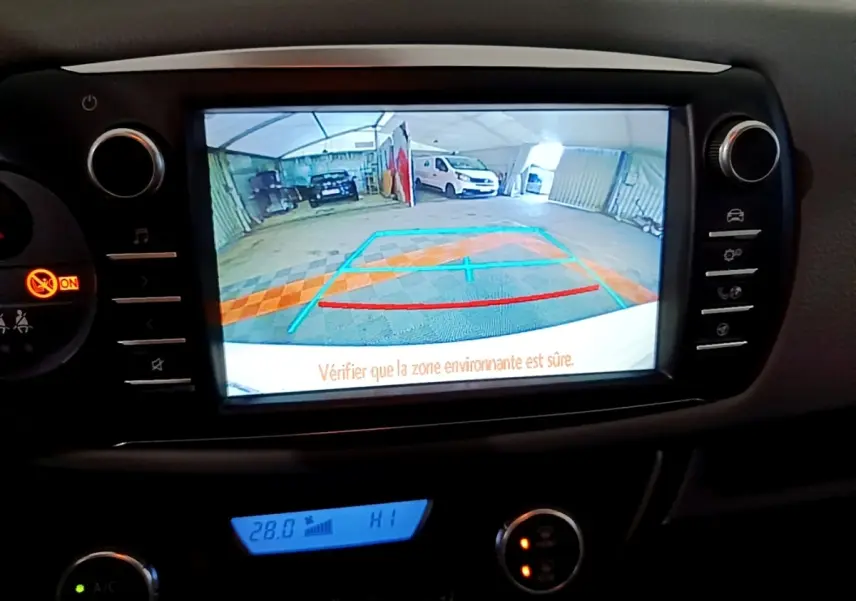 Écran tactile central affichant la caméra de recul d'une Toyota Yaris blanche, vue intérieure du tableau de bord.