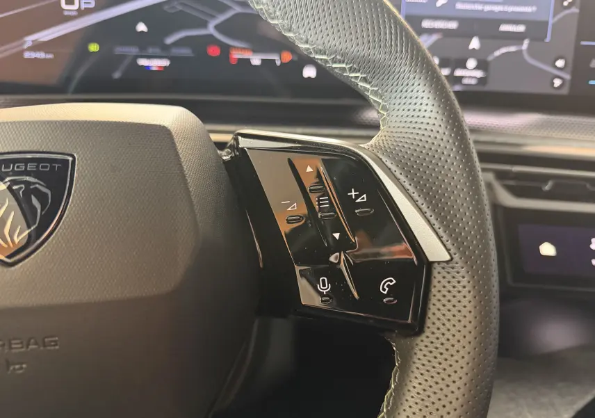 Gros plan sur le côté droit du volant en cuir perforé du Peugeot 3008 Hybrid 2025, avec commandes intégrées et tableau de bord numérique flou.