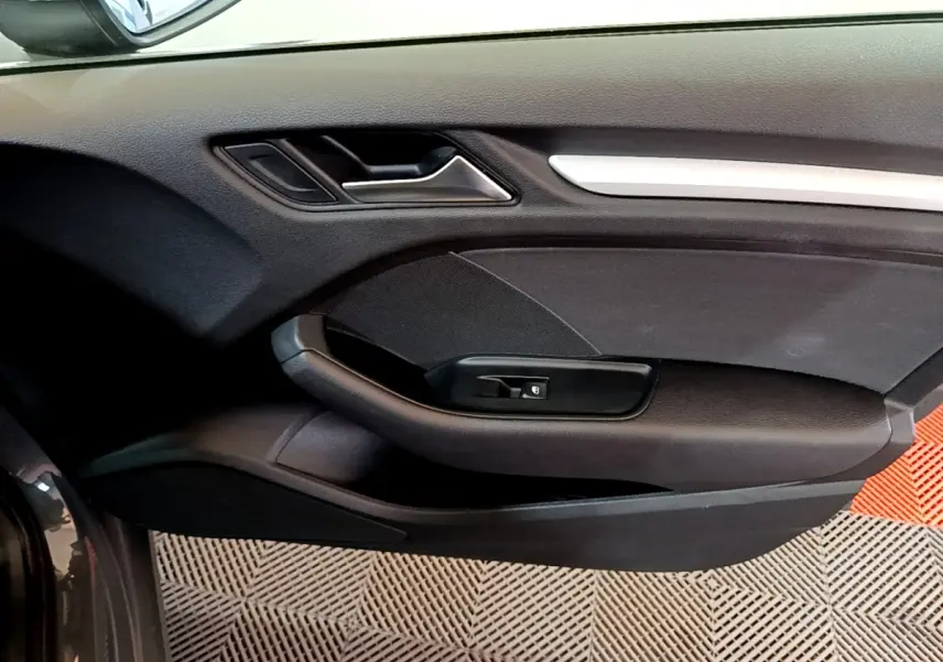 Vue intérieure côté gauche de la porte avant d'une Audi A3 gris foncé avec poignée argentée et commande de vitre électrique.