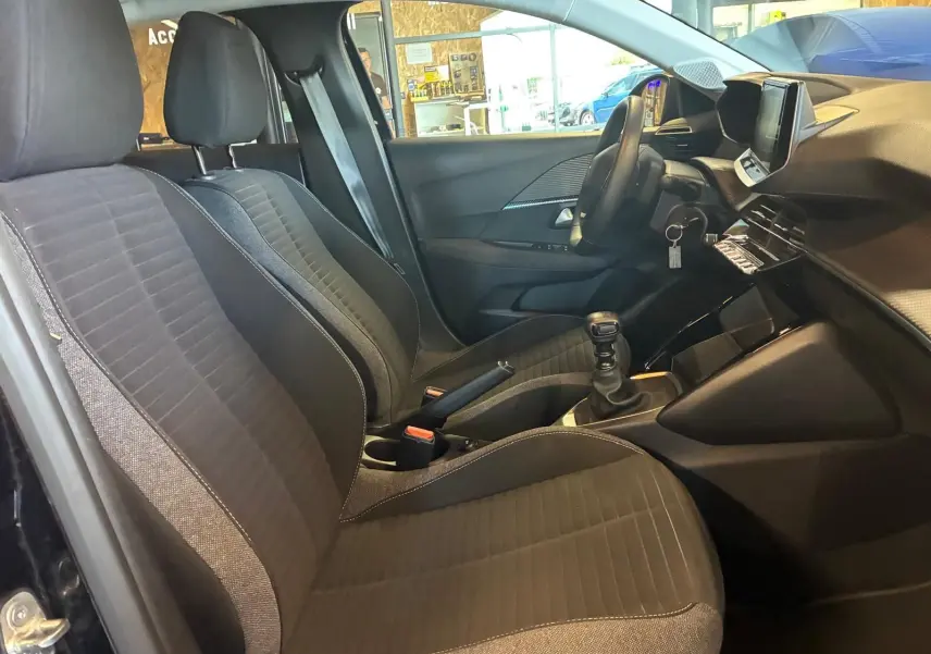 Vue intérieure côté conducteur de la Peugeot 208 noire, mettant en valeur les sièges en tissu et la console avec boîte manuelle.