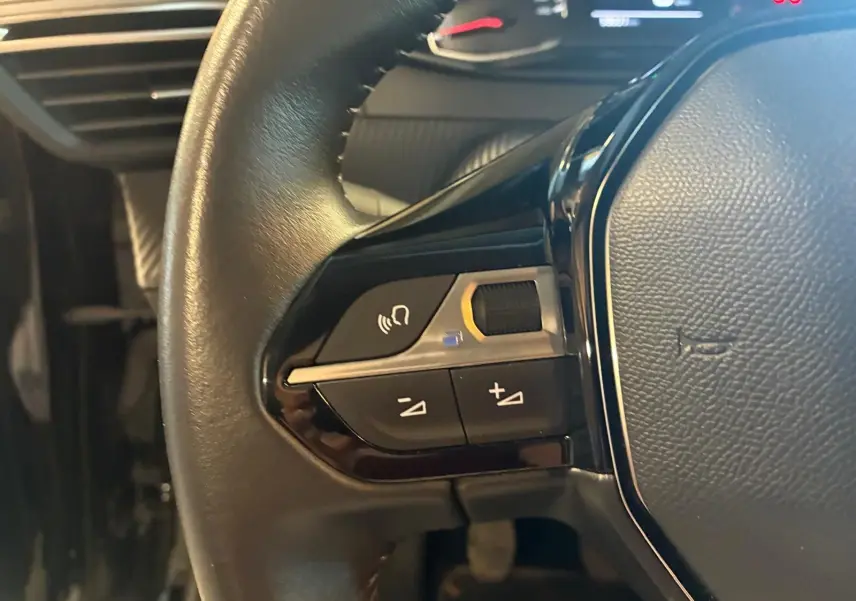 Gros plan sur les commandes gauche du volant noir de la Peugeot 208 1.5 BlueHDi 100 Style, tableau de bord en arrière-plan.