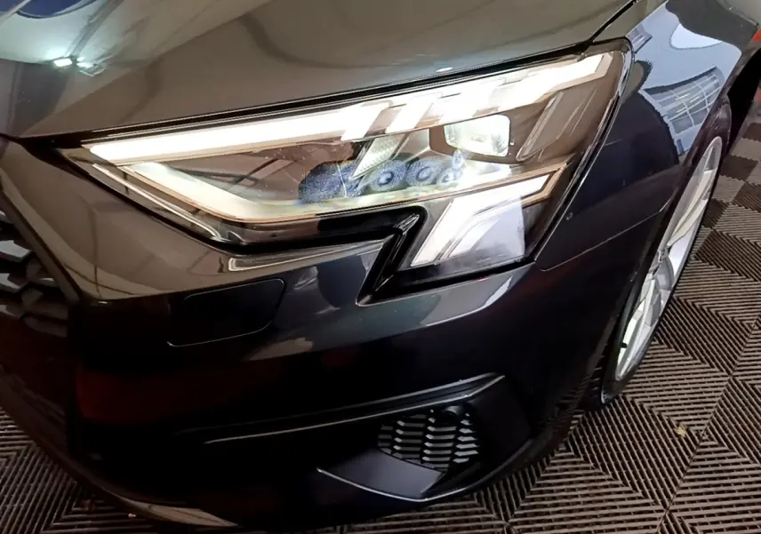Gros plan sur le phare avant droit d'une Audi A3 gris foncé avec détails LED et partie de la calandre visible.