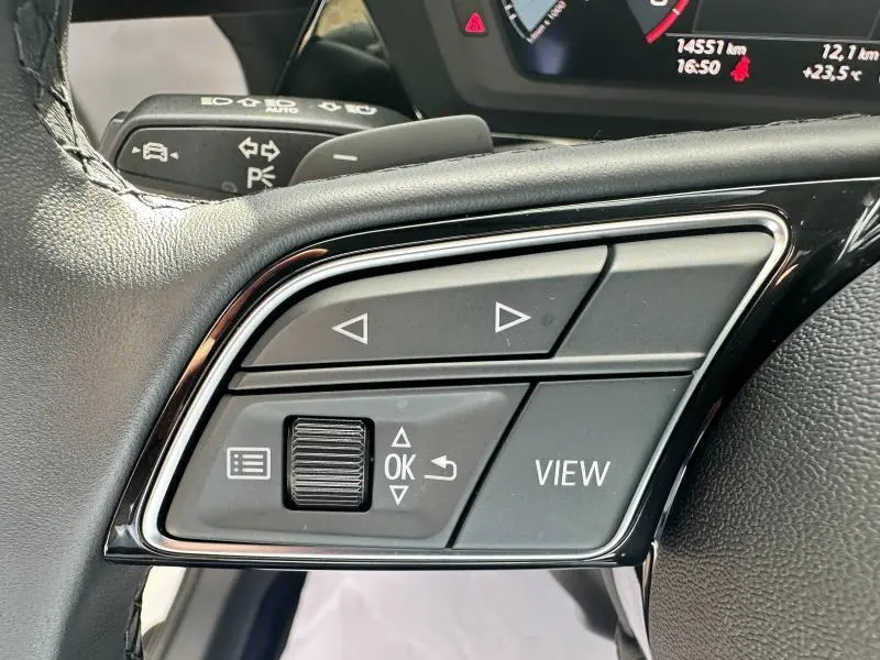 Gros plan sur les commandes multifonctions noires du volant cuir de l'Audi A3 Sportback 2024, avec tableau de bord partiellement visible.