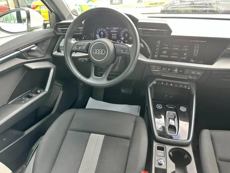 Vue intérieure avant de l'Audi A3 Sportback 2024, volant cuir multifonction, écran tactile et sièges tissu noir avec bande grise centrale.