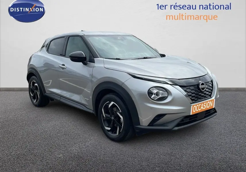 Nissan Juke hybride gris perle métal en 3/4 avant droit, avec jantes noires et calandre distinctive.