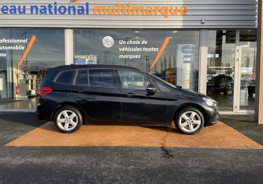 Profil côté gauche d'une BMW Série 2 Gran Tourer F46 noire, garée devant une concession multimarque en plein jour.