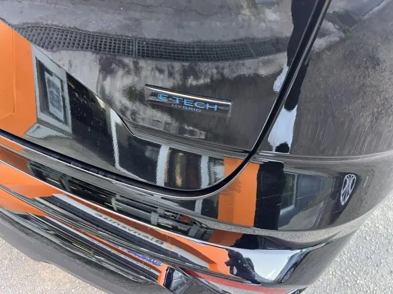 Gros plan sur le logo E-Tech Hybrid à l'arrière d'une Renault Austral noir étoilé, reflet d'un mur orange visible.