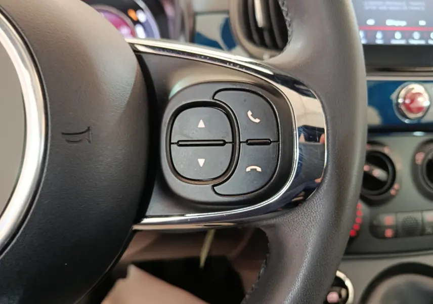 Gros plan sur les commandes du volant cuir noir de la Fiat 500 Hybrid bleu, avec tableau de bord et écran tactile en arrière-plan.