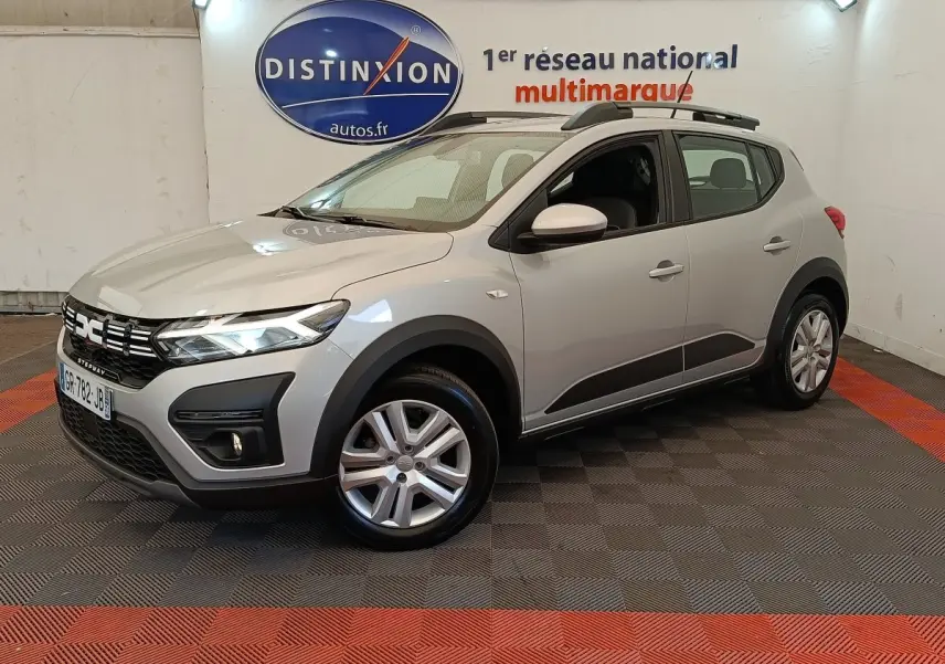 Vue 3/4 avant droit d'une Dacia Sandero Expression gris clair avec barres de toit et jantes alliage en showroom.