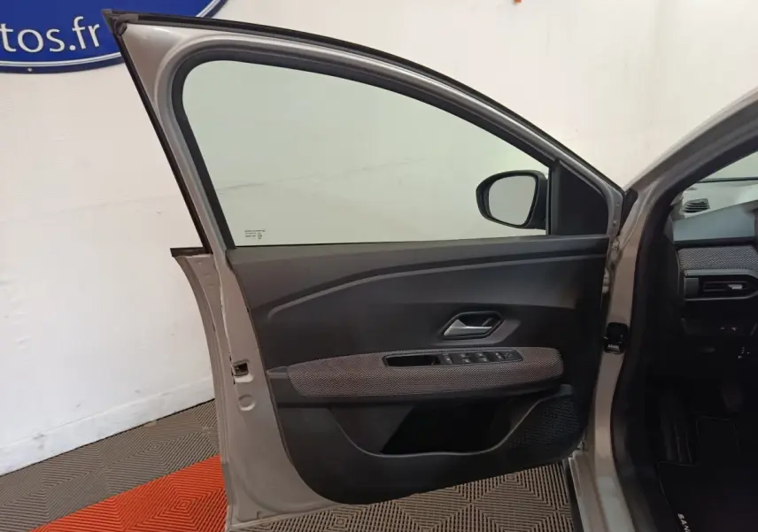 Porte avant gauche ouverte d'une Dacia Sandero gris clair 2023, montrant le panneau intérieur avec commandes électriques.