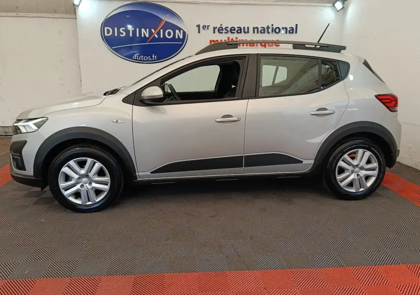 Vue de profil côté gauche d'une Dacia Sandero gris clair 2023 avec protections noires et barres de toit.