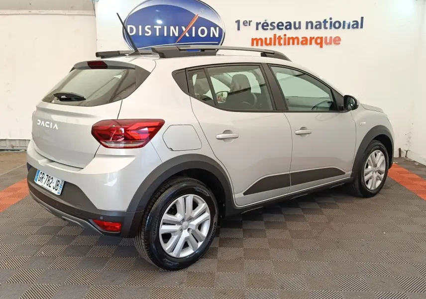 Vue 3/4 arrière droite d'une Dacia Sandero gris clair 2023 avec barres de toit et protections noires sur les bas de caisse.