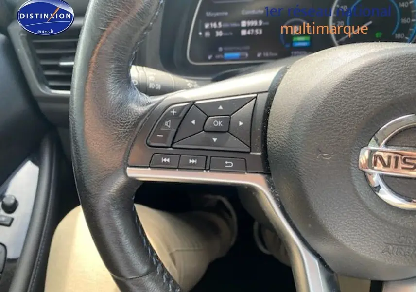 Gros plan sur le volant en cuir noir d'une Nissan Leaf électrique blanche nacrée, avec commandes intégrées et tableau de bord numérique visible.