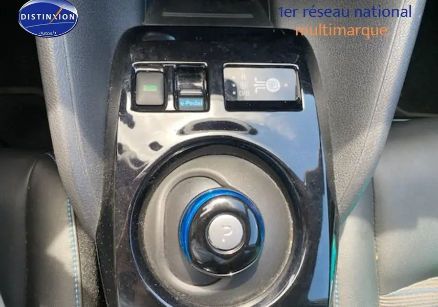 Gros plan sur la console centrale noire brillante avec sélecteur de vitesse rond et boutons de mode de conduite du Nissan Leaf blanc nacré.