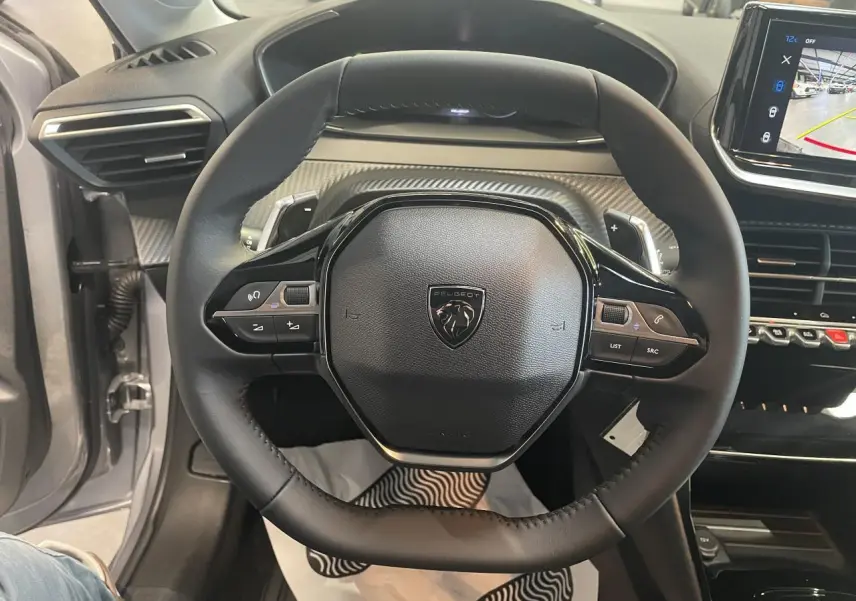 Vue rapprochée du volant cuir noir de la Peugeot 208 2025 avec commandes intégrées et tableau de bord carbone.