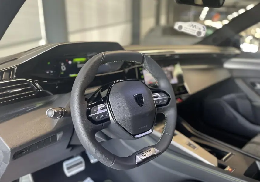 Volant cuir noir avec logo Peugeot et badge GT, planche de bord moderne et combiné numérique d'une 308 GT 2025.