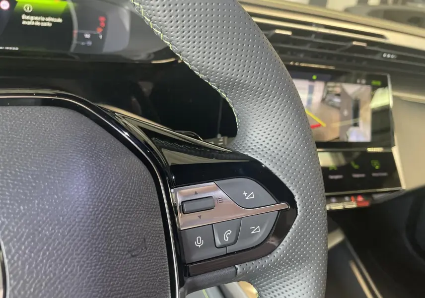 Gros plan sur le volant cuir perforé avec commandes intégrées et écran numérique du tableau de bord de la Peugeot 308 blanche.