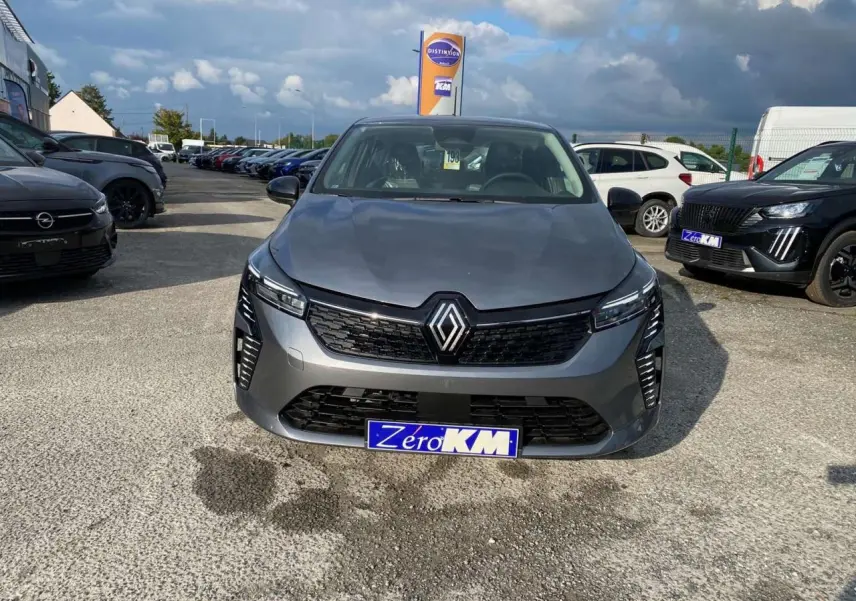 Vue frontale d'une Renault Clio 2025 gris schiste avec calandre noire et plaque "Zéro KM" sur un parking.