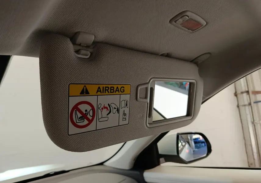 Détail intérieur du pare-soleil côté conducteur beige avec miroir et avertissement airbag, rétroviseur extérieur visible.