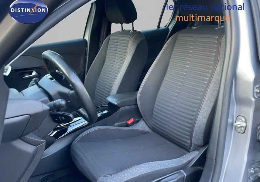 Vue intérieure côté conducteur de la Peugeot 208 électrique gris Artense, sièges tissu gris avec surpiqûres rouges.