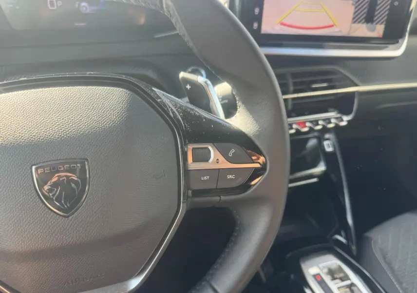 Gros plan sur le volant cuir noir de la Peugeot 2008 Hybrid 145, avec commandes intégrées et écran tactile en arrière-plan.