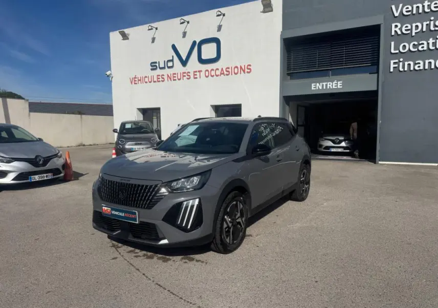 Peugeot 2008 Hybrid 145 gris vue 3/4 avant droit, jantes alliage 17 pouces, calandre noire distinctive, stationné devant un garage.
