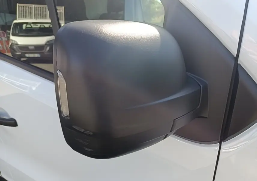 Gros plan sur le rétroviseur noir côté conducteur du Renault Trafic Fourgon blanc, avec reflet d’un autre véhicule en arrière-plan.