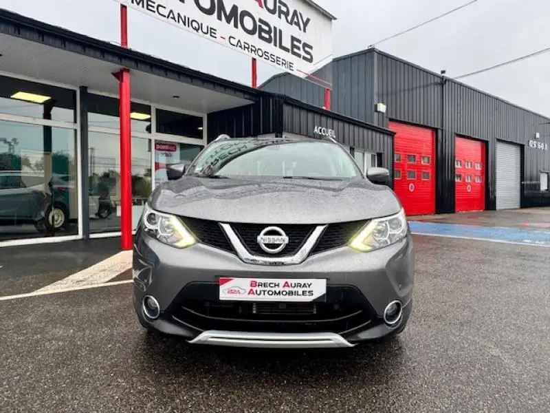 Vue frontale d'un Nissan Qashqai gris Squale avec phares allumés devant un garage automobile.