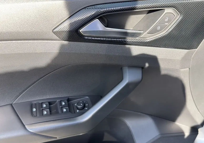 Vue rapprochée de la porte côté gauche du Volkswagen T-Cross 2025 en tissu noir avec commandes électriques et poignée grise.