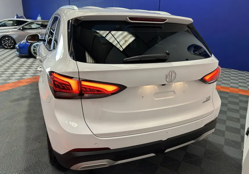 Vue 3/4 arrière droite du MG ZS 1.5 Hybrid+ 197 Luxury en blanc Dover avec feux arrière allumés dans un showroom.