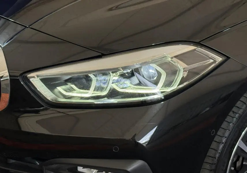 Gros plan sur le phare avant droit allumé d’une BMW Série 1 116d noire, avec partie de la roue visible.