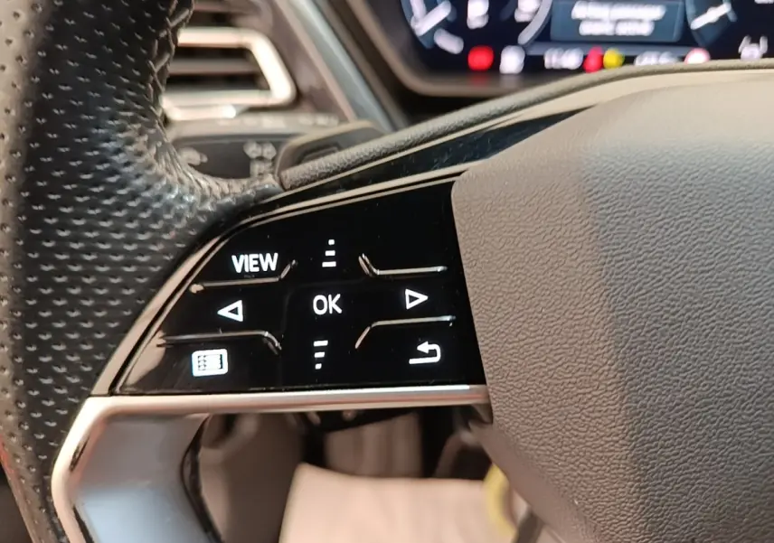 Gros plan sur les commandes gauche du volant cuir perforé de l'Audi Q4 E-TRON Sportback noir, avec affichage numérique flou en arrière-plan.