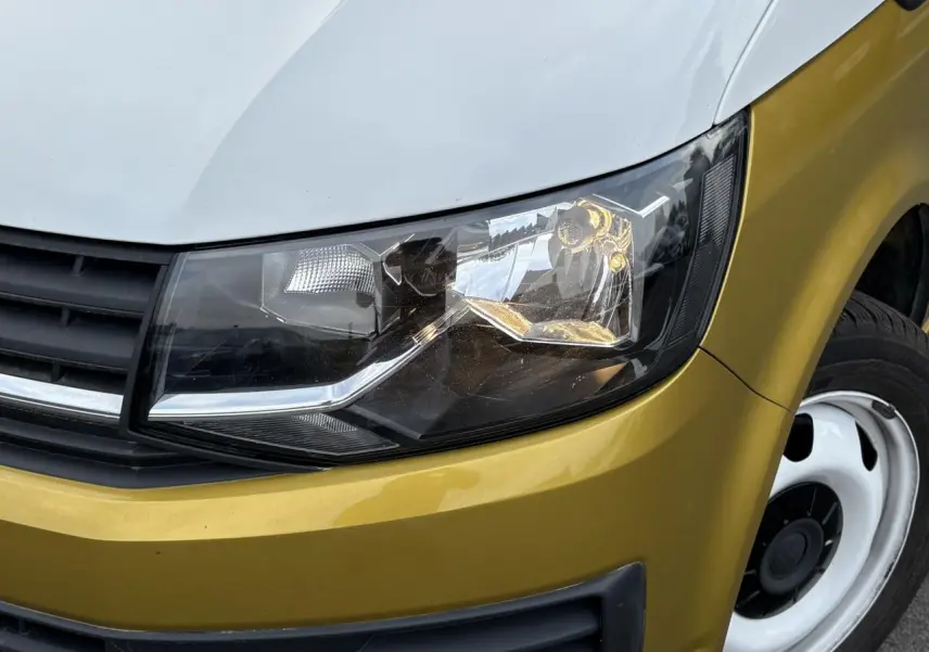 Gros plan sur le phare avant droit du Volkswagen Transporter 2.0 TDI jaune et blanc, mettant en valeur la calandre noire et la jante acier.