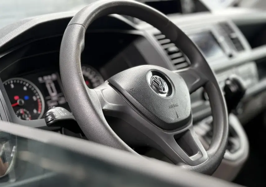 Vue rapprochée du volant noir du Volkswagen Transporter 2017, avec tableau de bord et commandes visibles en arrière-plan.