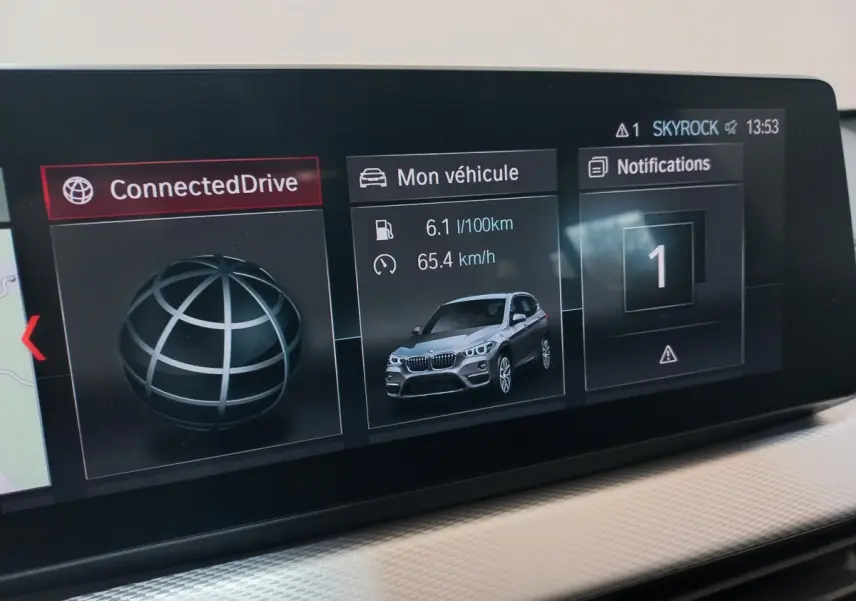 Écran tactile intérieur montrant la consommation et la vitesse d'un BMW X1 noir en vue 3/4 avant.