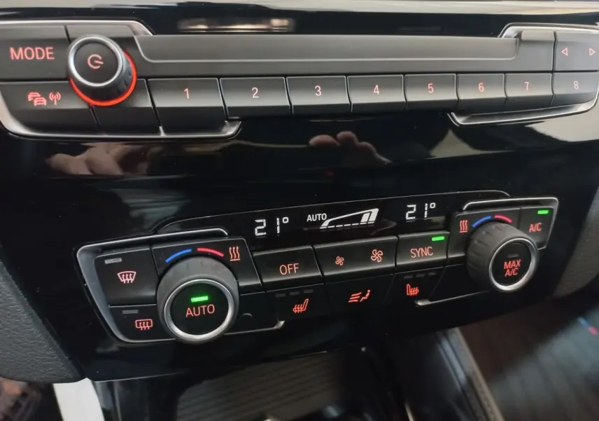 Gros plan sur la console de climatisation digitale de la BMW X1 noire, avec réglages à 21°C et boutons rétroéclairés rouges.