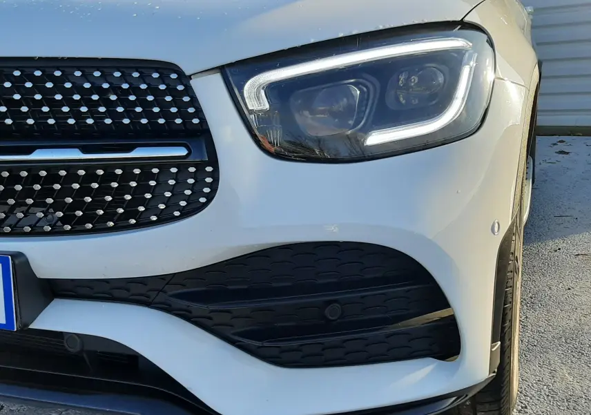 Gros plan sur l'avant droit d'un Mercedes GLC Coupé blanc avec calandre AMG et phares LED allumés.