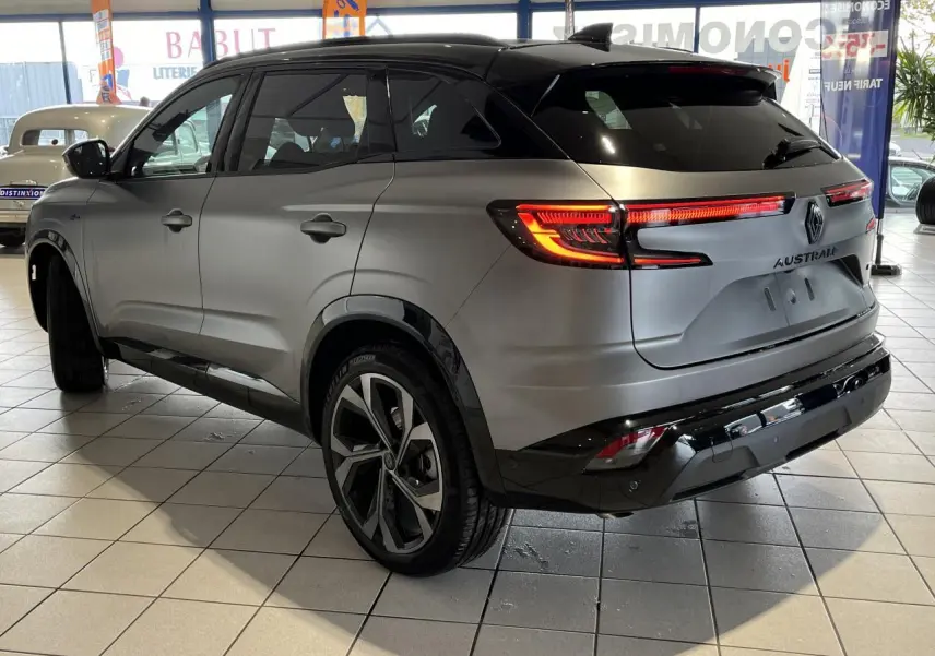 Vue 3/4 arrière droite du Renault Austral gris avec toit noir étoilé et feux arrière LED allumés en intérieur showroom.