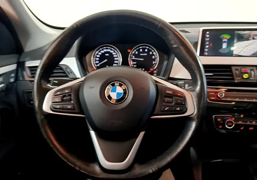 Vue intérieure centrée sur le volant cuir noir d’une BMW X1 2020 avec tableau de bord et écran tactile visible à droite.