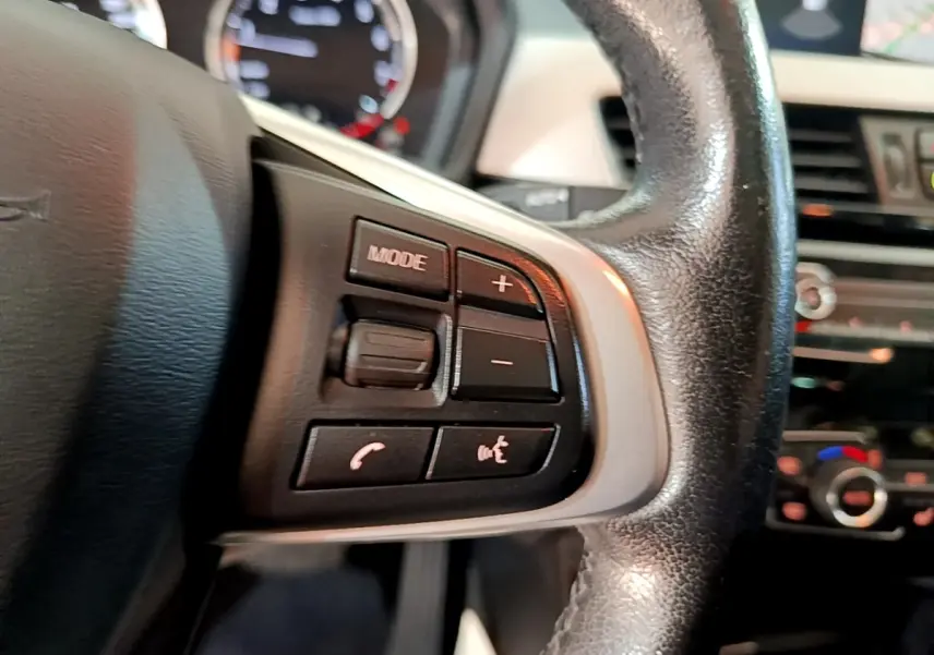 Gros plan sur les commandes multimédias au volant cuir noir d'une BMW X1 sDrive20i 2020, intérieur visible flou.